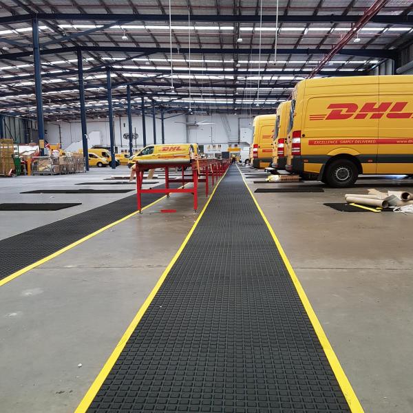 Yellow DHL vans back onto a walkway fitted with the Air Grid anti fatigue mat. The mat is black with a yellow safety border.