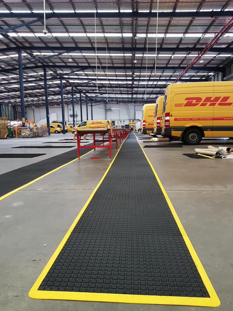 Yellow DHL vans back onto a walkway fitted with the Air Grid anti fatigue mat. The mat is black with a yellow safety border.