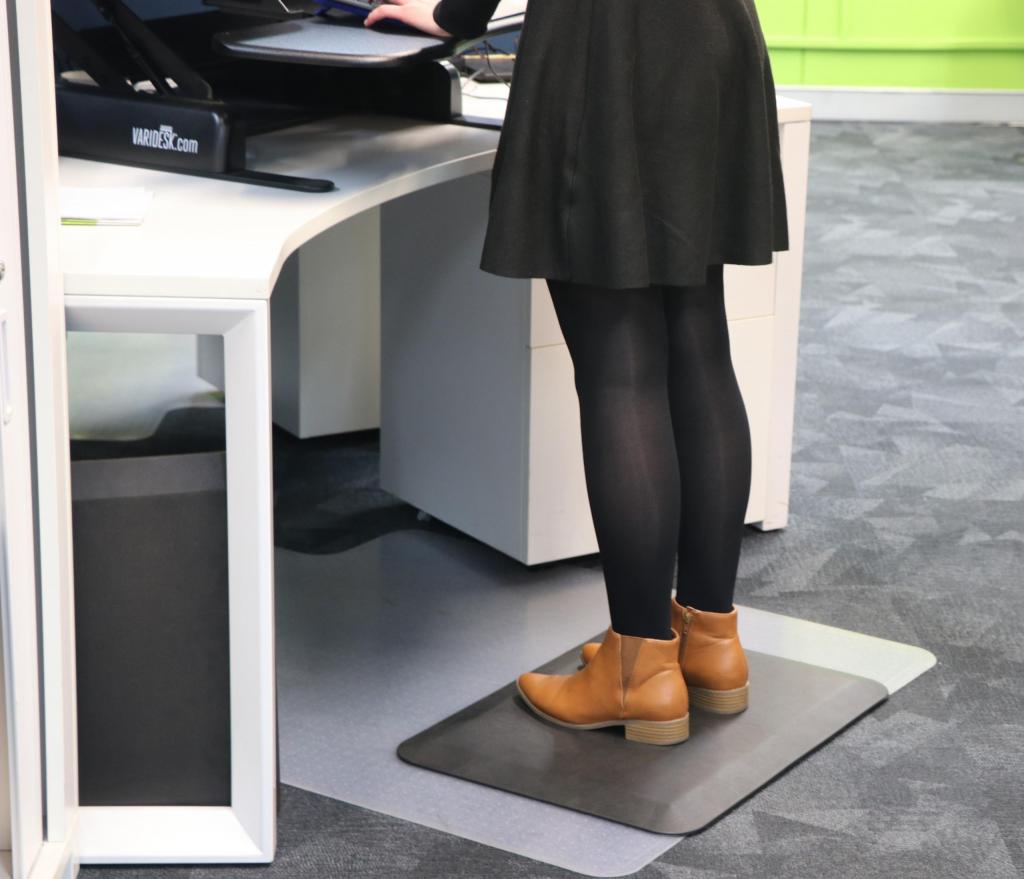 a woman in a black skirt, black tights and brown boots stands on an anti fatigue mat (the comfort stand plus) while working at a stand up desk.