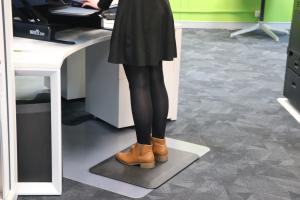 a woman in a black skirt, black tights and brown boots stands on the comfort stand plus (an anti fatigue mat) while working at a stand up desk.