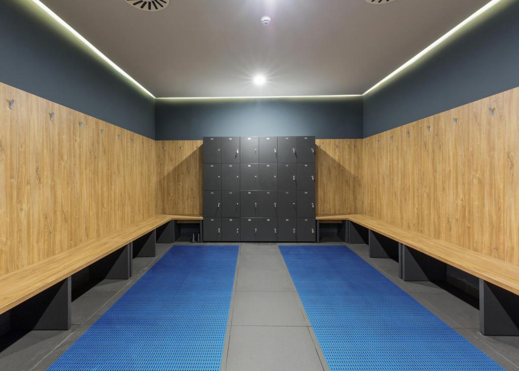 A locker room with wooden benches around the outside and a block of lockers at the end. The floor is tiled with Sure Grippa non slip matting laid in two strips covering most of the floor except the centre.