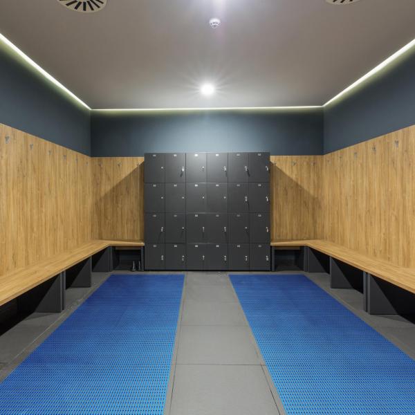 A locker room with wooden benches around the outside and a block of lockers at the end. The floor is tiled with Sure Grippa non slip matting laid in two strips covering most of the floor except the centre.