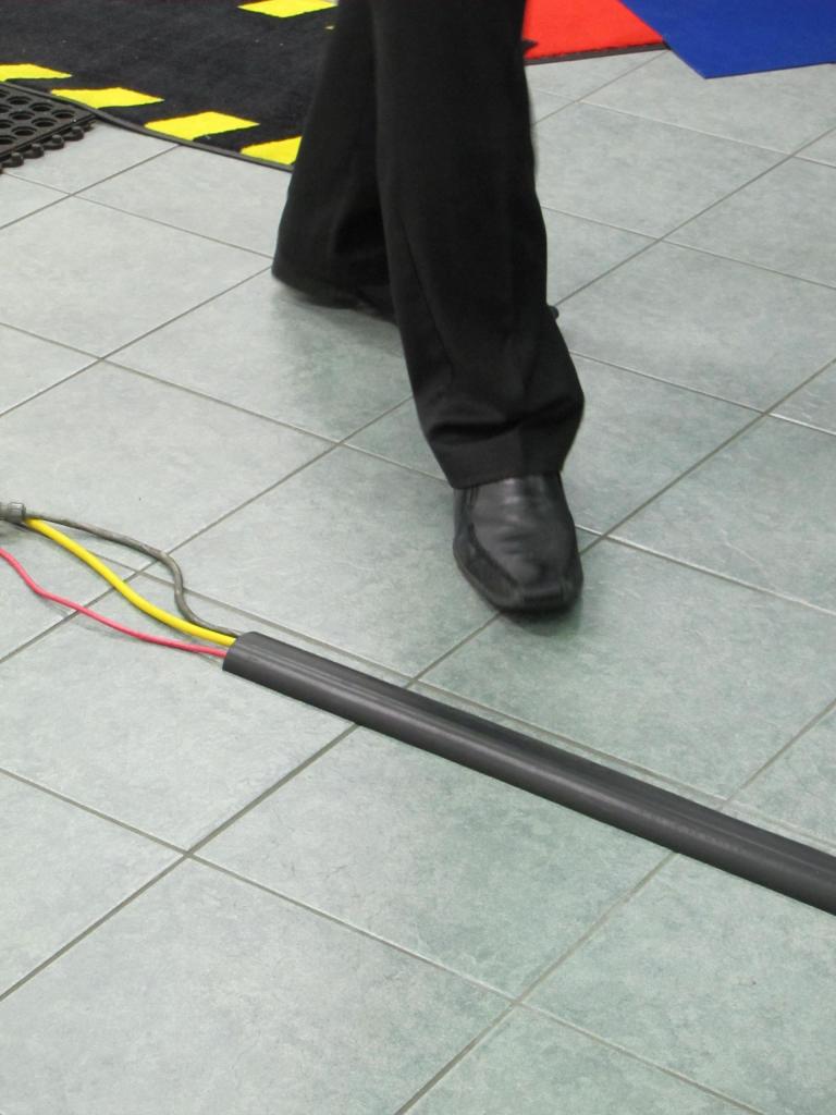 A cable protector is laid on a pavement over 3 wires. A person is bout to step over the wires safely thanks to the cable protector.