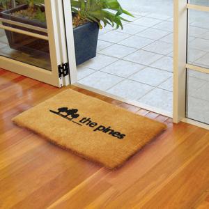 a coir entrance mat with black branding "The Pines" shows 3 trees sketched. The mat is laid on a wooden floor.