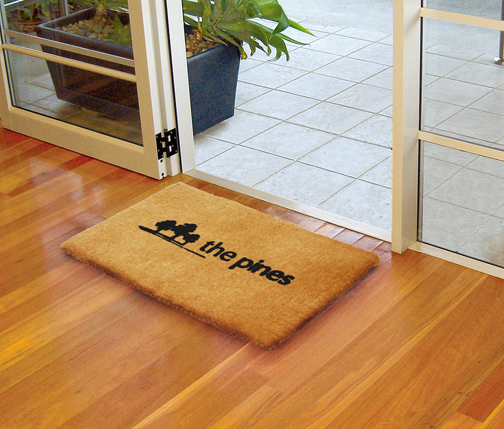 a coir entrance mat with black branding "The Pines" shows 3 trees sketched. The mat is laid on a wooden floor.