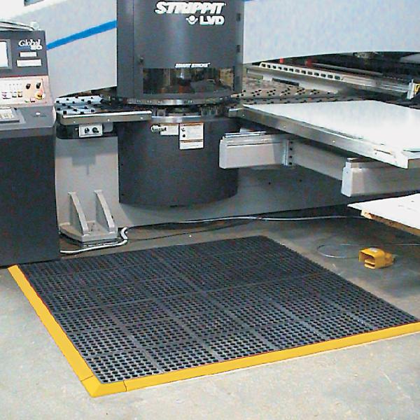 the comfort link grit top is laid in front of a factory machine. The machine is large with multiple layers and the mat is interlocking with a yellow safety border.