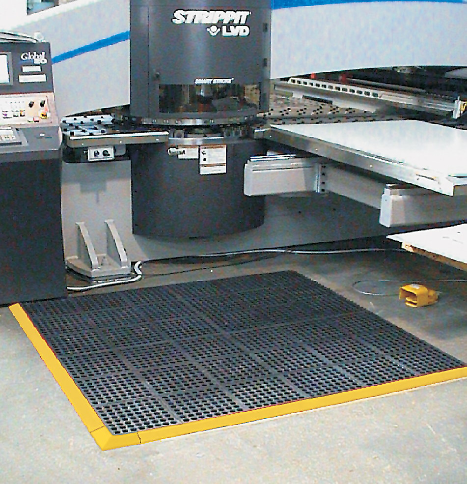 the comfort link grit top is laid in front of a factory machine. The machine is large with multiple layers and the mat is interlocking with a yellow safety border.
