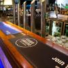 A bar with beer taps, glasses and various drinks. Printed Bar runners are placed along the front of the bar top. One is printed with the logo of a hotel and the other is printed with a coffee roaster logo.