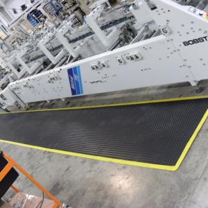 The ergo tread anti fatigue mat is laid in front of a BOBST machine. The machine is grey with dials and switches along the front. The mat is black with a yellow safety border.