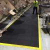 The comfort link is laid down a warehouse aisle where cardboard boxes are being loaded from conveyer belts. A man is working on the black anti fatigue mat.