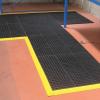 Custom shape comfort link modular mats are laid at a work bench. The bench is blue on a red floor and the mat is black with a yellow safety border.