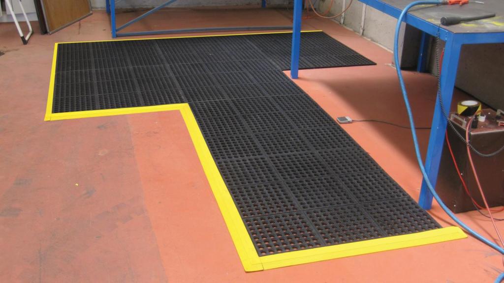 Custom shape comfort link modular mats are laid at a work bench. The bench is blue on a red floor and the mat is black with a yellow safety border.