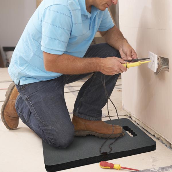 A sparky uses the knee saver while he works on a plug socket. He is holding yellow and black tools and there is a red screwdriver nearby.