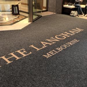 The entrance to The Langham Melbourne is shown with a round doorway and a large entrance mat. The mat is printed with the name of the hotel in gold letters. A group of people are sitting in a cove to the right of the mat.