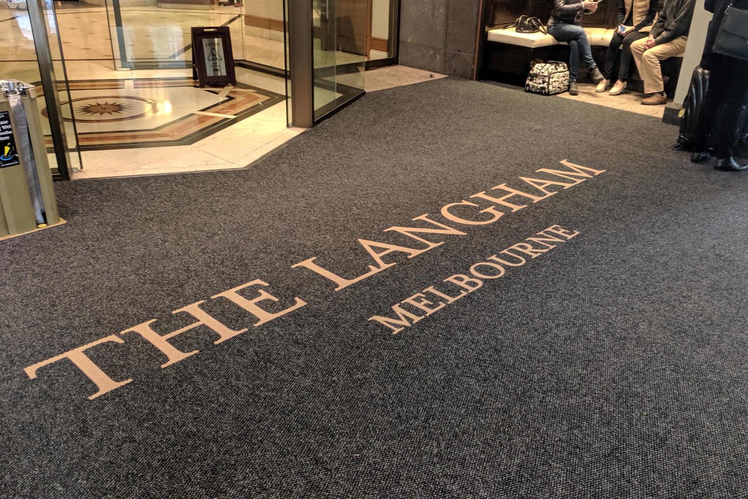 The entrance to The Langham Melbourne is shown with a round doorway and a large entrance mat. The mat is printed with the name of the hotel in gold letters. A group of people are sitting in a cove to the right of the mat.