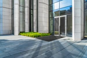 The Opti-Scrape entrance mat is laid in front of office building. The building is glass and stone and the sun can be seen shining through the glass.