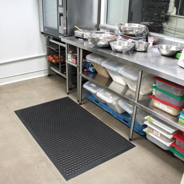 the cushion ease anti fatigue non slip mat is laid in a busy commercial kitchen. The mat is black with holes for drainage.