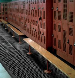 A locker room containing red metal lockers with individual padlocks, wooden benches and a comfort link non slip anti fatigue mat is pictured.