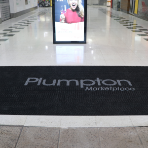 The entrance to a shopping centre is fitted with a non slip entrance mat printed with the words 'Plumpton Marketplace'