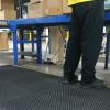 a pair of legs in black trousers, black workbooks and a yellow high visibility jacket is standing on a heavy duty anti fatigue comfort mat. There is a table stacked with boxes in front of them and a conveyer belt to their right.