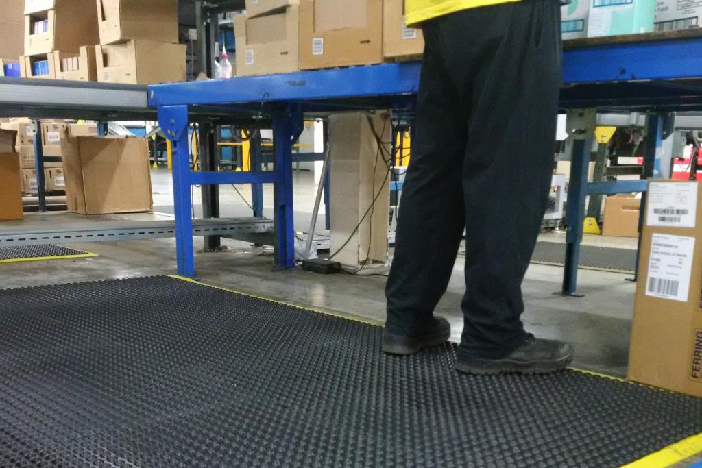 a pair of legs in black trousers, black workbooks and a yellow high visibility jacket is standing on a heavy duty anti fatigue comfort mat. There is a table stacked with boxes in front of them and a conveyer belt to their right.
