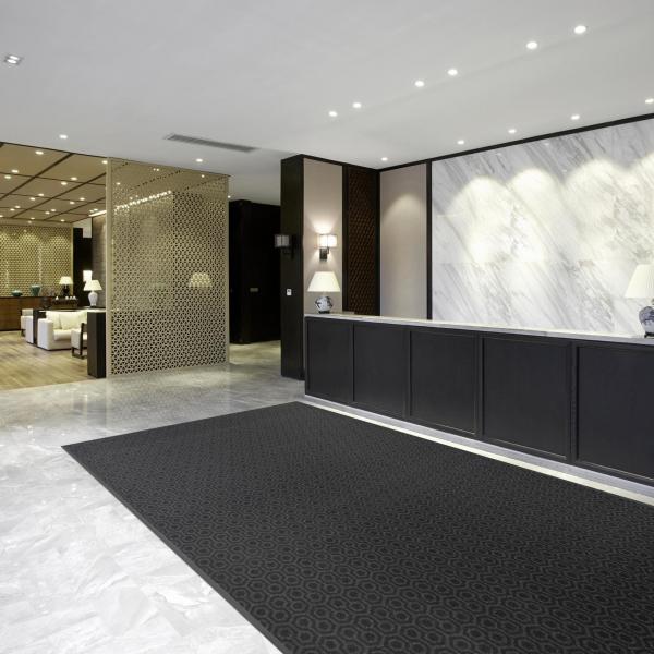 The Glamour Step entrance mat is laid in the reception area of a hotel. The reception has marbled white walls and floors with a black counter, and a lobby with sofas can be seen in the background.