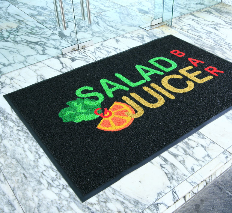 An entrance mat - the Vinyl Loop - is laid on a marble floor outside a glass door with metal handles. The mat is printed with the branding for Salad & Juice Bar in bright colours