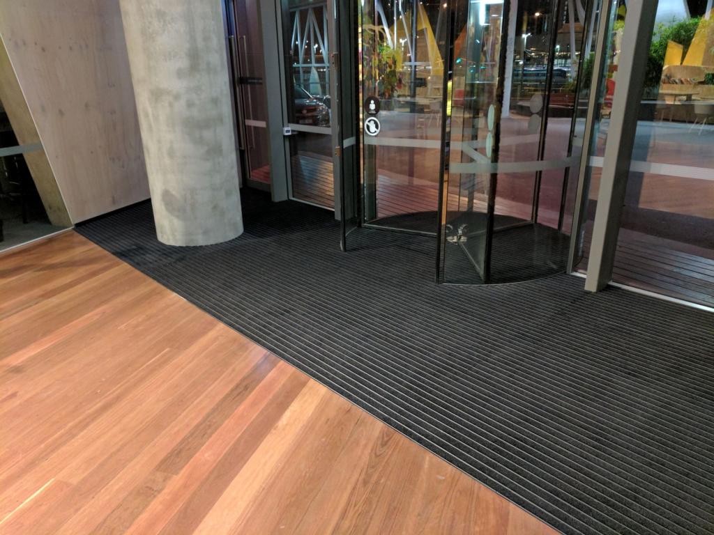 A rotating glass door divides an entranceway from an outside courtyard. The floor inside is wood style with a black entrance mat - the Esteem Ribbed II.