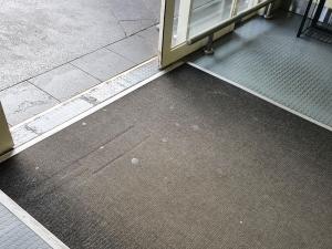 An old entrance mat with chewing gum stains is fitted into plastic flooring in an entranceway