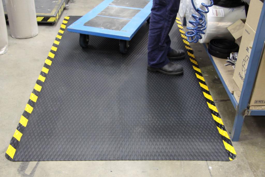 A pair of legs in navy trousers and black work boots is standing on the dura step anti fatigue mat. A small trolley is also on the mat, which has a black diamond patterned surface and a yellow and black border.