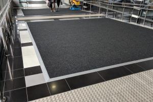 A large square entrance mat is fitted with silver metal ramped edges into a monochrome tiled floor. A children's car ride is seen in the background along with an elderly man with a walking stick