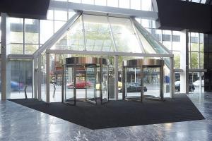 A trapezoid-shaped entrance mat is laid around two rotating glass doors. The floor beyond its is marbled-style tiles and the street outside has pavement and a busy road.
