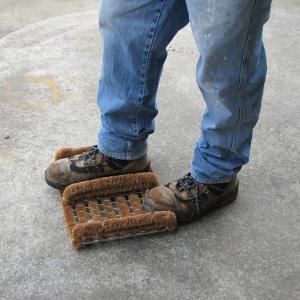 A man scrapes his workbook on the Boot Scraper. He is wearing blue jeans and is standing on a concrete floor.
