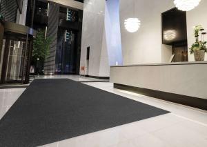 A sleek, modern building lobby featuring a large dark floor mat placed over glossy white tiles. The space includes tall ceilings, stylish lighting fixtures, and a reception desk, creating a clean and professional entrance area.