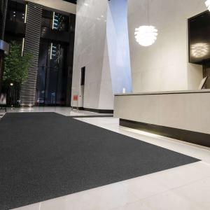 A sleek, modern building lobby featuring a large dark floor mat placed over glossy white tiles. The space includes tall ceilings, stylish lighting fixtures, and a reception desk, creating a clean and professional entrance area.