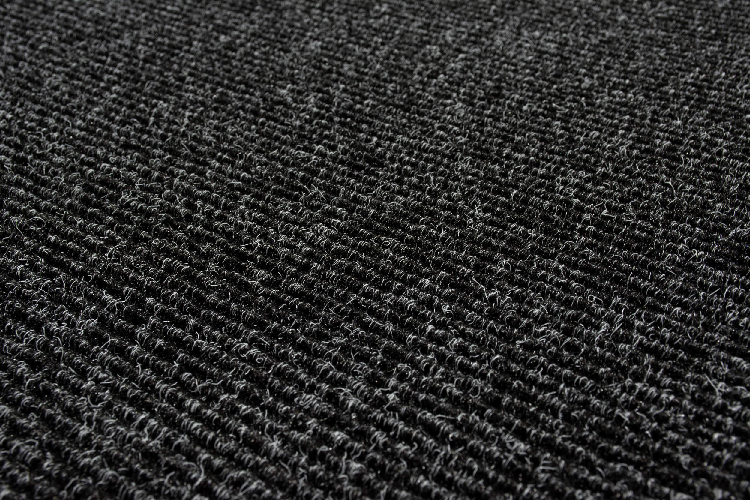 A close-up view of a dark gray textured carpet surface, featuring tightly looped fibers and a ribbed pattern.