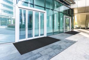 A large black entrance mats placed outside a modern commercial building, creating a clean and safe entryway. The mats help capture dirt and moisture before visitors step inside, maintaining a polished and professional look.