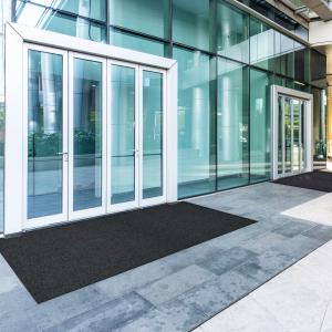 A large black entrance mats placed outside a modern commercial building, creating a clean and safe entryway. The mats help capture dirt and moisture before visitors step inside, maintaining a polished and professional look.